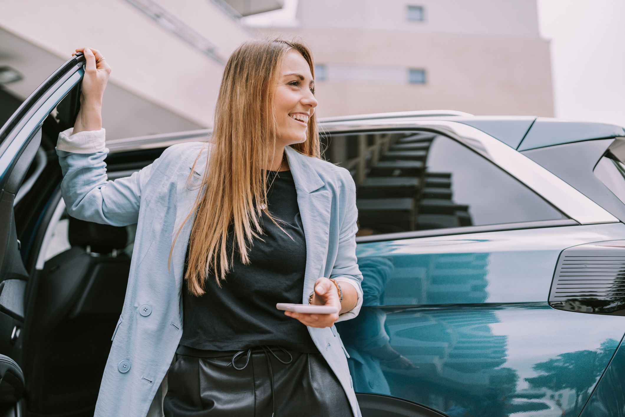 Junge Frau steigt lächelnd aus einem modernen Auto aus und hält ein Smartphone in der Hand